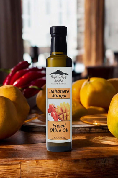 Bottle of Habanero Mango Fused Olive Oil surrounded by fresh fruits and peppers on a wooden table.