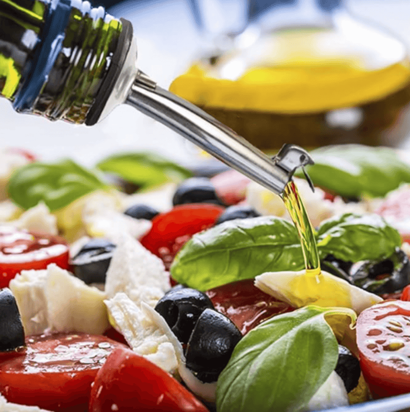 Stainless steel pour spout dispensing olive oil over a fresh salad with tomatoes, mozzarella, and basil.